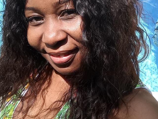 LucyLina69's face, smiling with dark curly hair, brown eyes, and a cute nose, wearing a green patterned top, sunlit, showing her shiny brown skin and red lips.