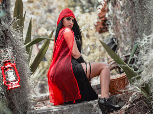 MartinaMendezz mit langem, schwarzem Haar, roter Kapuze, durchsichtigem roten Umhang, schwarzem Kleid, Strümpfchenband, schwarzen Absätzen, sitzend auf einem Felsen, wobei ihr Hintern zu sehen ist, in einer wüstenähnlichen Umgebung mit einer Laterne..