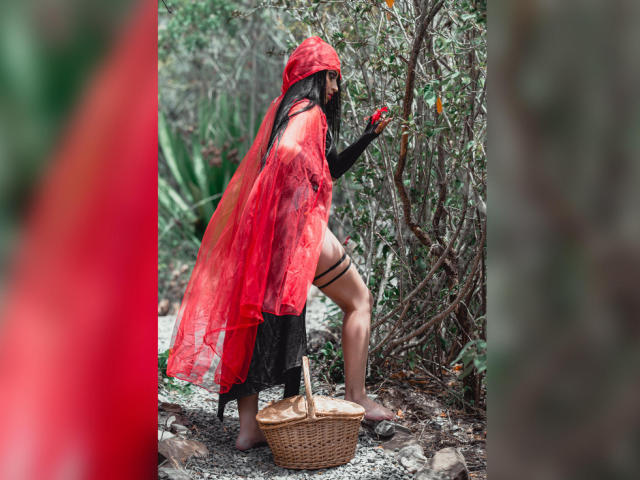 MartinaMendezz in een rode, doorschijnende, bloedkleurige mantel, een zwarte top met lange mouwen en zwarte dijbanden, met een rood object in haar hand, en staand op blote voeten naast een rieten mand..