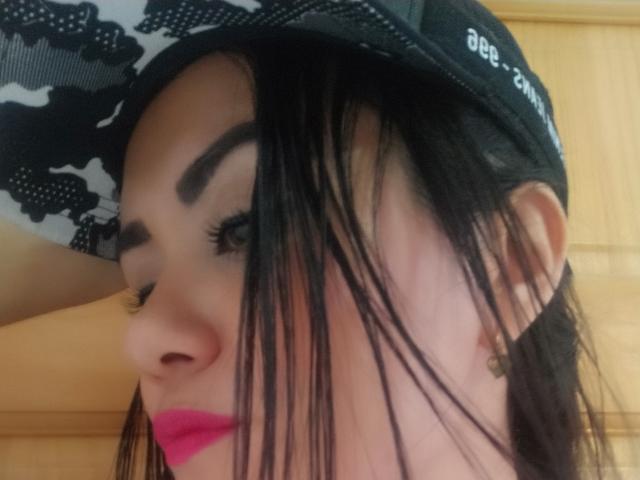 XBellaEvaX's face with black hair, pink lipstick, and thick eyebrows, wearing a black and white camouflage baseball cap, showing off long eyelashes and a cute nose.