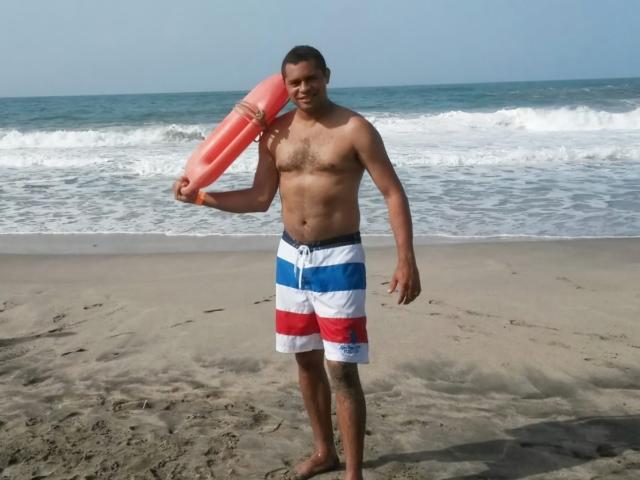 Shirtless LatinXBlackHard dude with a medium build and dark skin stands on the beach, holding a pink boogie board in his right hand. He's wearing red, white, and blue shorts.