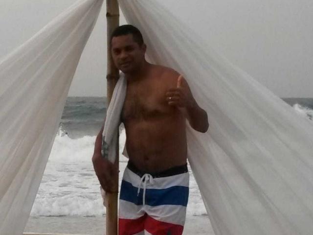 LatinXBlackHard, sin camisa, con una toalla sobre un hombro, usando shorts de baño rojos, blancos y azules, de pie debajo de cortinas blancas, haciendo un gesto de pulgar hacia arriba. Océano y olas en el fondo..