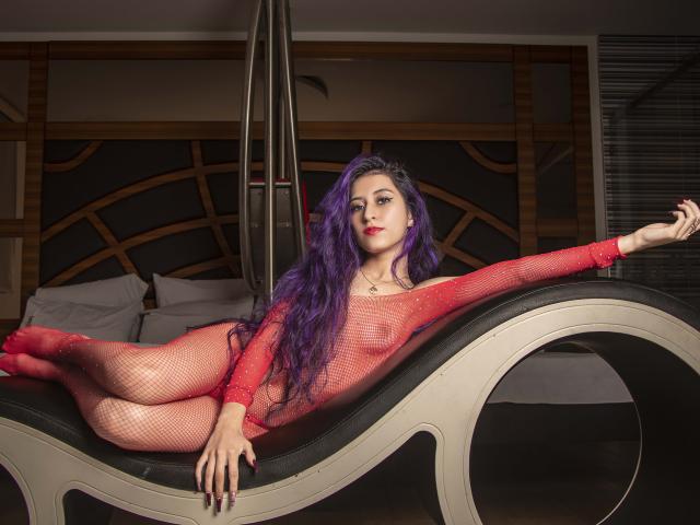 JuliaJobs with long purple hair, red fishnet bodysuit, and red socks, laying on a black and white curved chaise, exposing her tits and pussy through the sheer fabric.