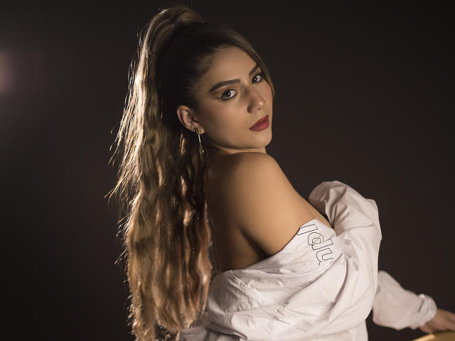 SusiAndMat con lunghi capelli castano ondulati raccolti in una coda di cavallo, camicia bianca a spalle scoperte, rossetto rosso, che guarda oltre la spalla, sfondo scuro, luce soffusa, orecchini, espressione delicata..