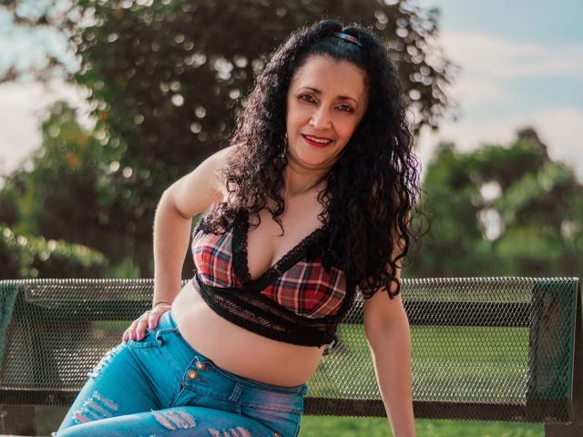 TitaNaughty with curly black hair, red plaid bralette showing her tits, blue ripped jeans, and a smile, leaning on a green metal bench in a sunny park.