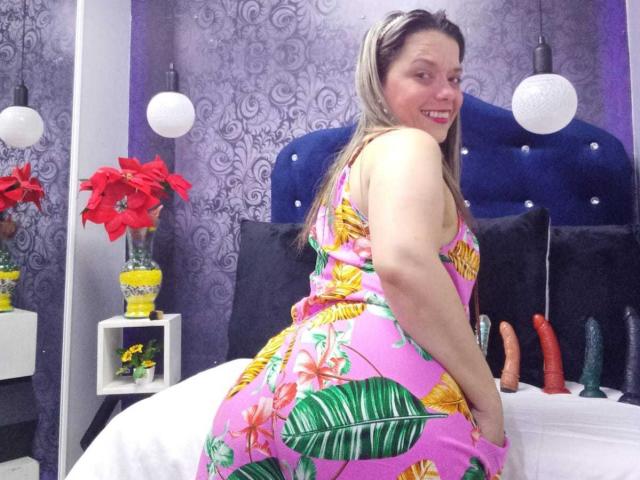 BiancaWild with pink floral dress, large ass, and blonde hair, smiling at camera, kneeling on bed with colorful dildo collection in background. Sheer pink dress highlights her curvy body.