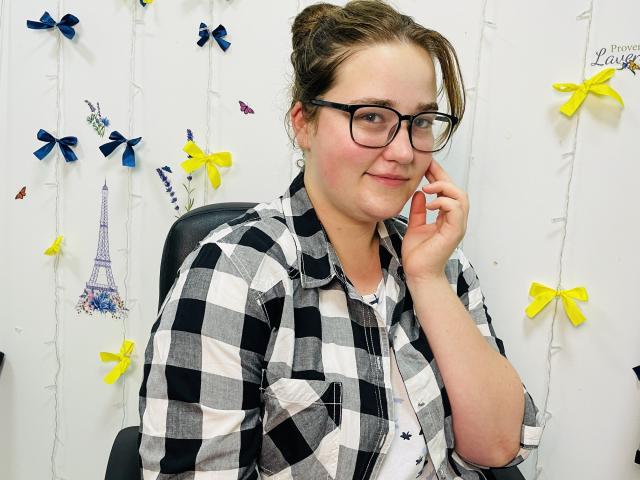 DaniellaGraceZZ with glasses, checking phone, wearing black-and-white checkered shirt, yellow and blue bows in background, slight smile, brown hair in bun, white wall, black chair.