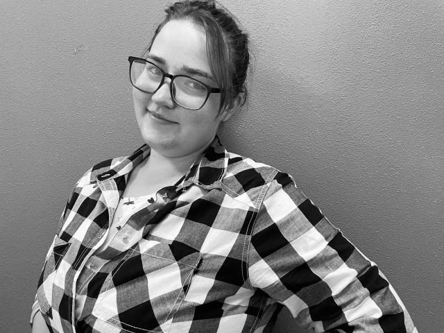 DaniellaGraceZZ has glasses, a checkered shirt, and a slight smile. She's leaning against a wall, looking relaxed with her hands behind her back. Her tits are small.