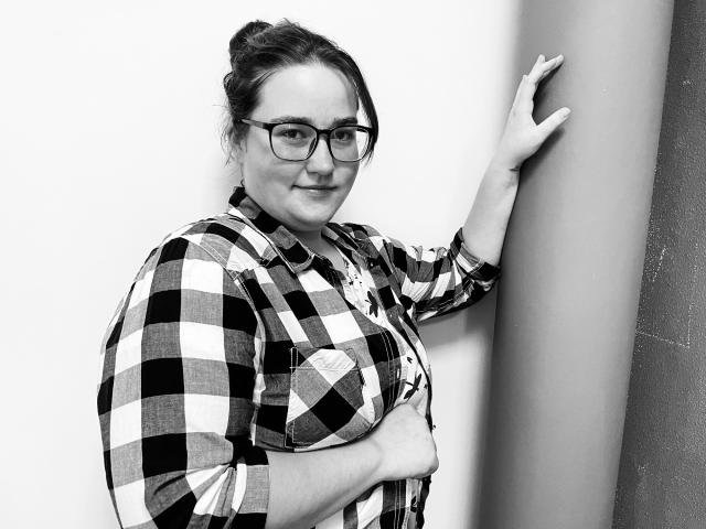 DaniellaGraceZZ, di taglia comoda e con gli occhiali, indossa una camicia a quadri, è appoggiata a un muro, con una mano che lo tocca, sorride leggermente, tenendo il seno con l'altra mano. Foto in bianco e nero..