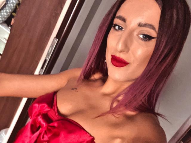 MissAlice69 met paars haar, rode lippenstift en zwarte eyeliner, draagt een rode satijnen jurk, toont haar borsten, kijkt naar de camera, in een slaapkamer..