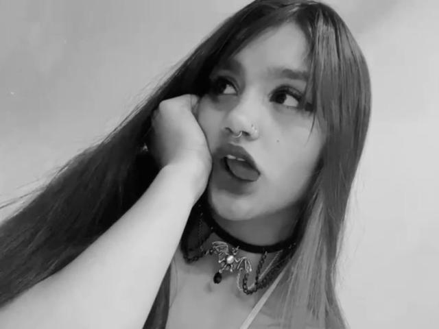 Gaby Durand mit langen Haaren, Nasenring und Choker, Hand am Kinn, Zunge heraus, blickt nach oben. Ihre Brüste sind teilweise sichtbar. Sie ist ein süßes, sexy Mädchen..