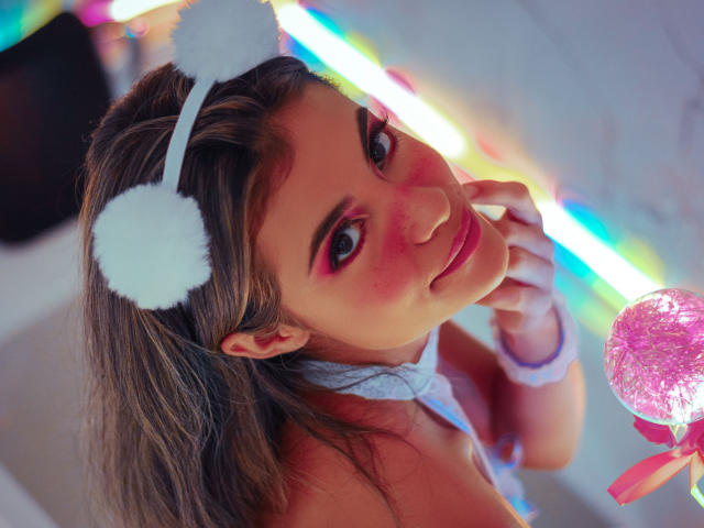 AbbyMoont con trucco occhi rosa, fascia bianca e soffice, e nastro grigio al collo, che guarda verso l'alto, con un dito vicino alla bocca, una sfera glitter rosa a destra e una striscia LED arcobaleno sullo sfondo..