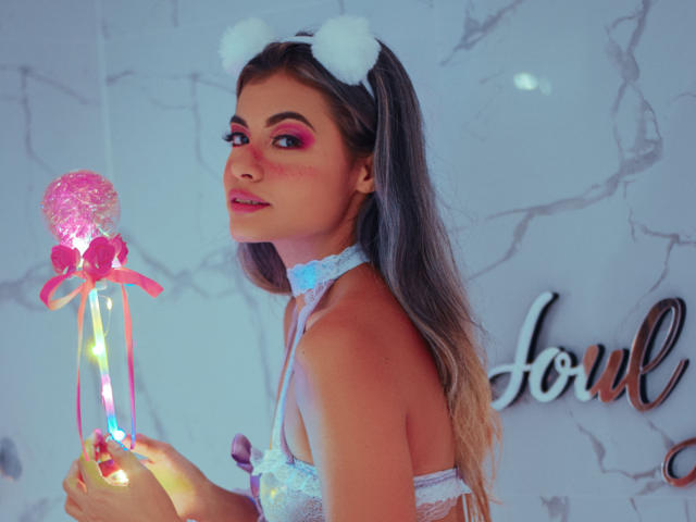 AbbyMoont with white furry cat ears, pink eye makeup, and a blue choker, holding a pink heart lollipop with a ribbon, wearing lace lingerie, showing her tits.