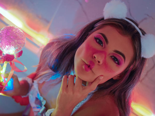 AbbyMoont, com sombra rosa, orelhas brancas e volumosas, e uma mão perto do rosto, exibindo sua bola rosa brilhante com um laço. Ela está corada com uma expressão adorável..