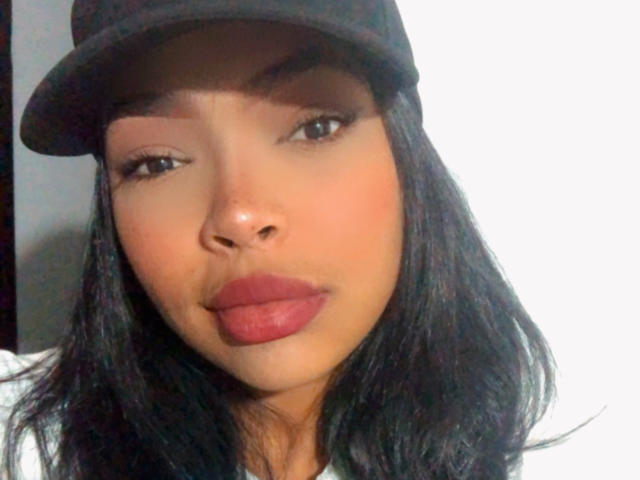 VickyGreys has dark brown hair, wearing a black cap, with red lipstick and a slight pout, showing a smooth, medium brown complexion, and small nose.