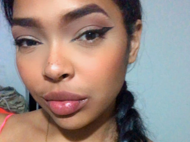 VickyGreys with thick eyebrows, black eyeliner wings, pinkish-brown lipstick, and a slight pout, her dark hair in a braid, wearing a pink top, against a plain background.