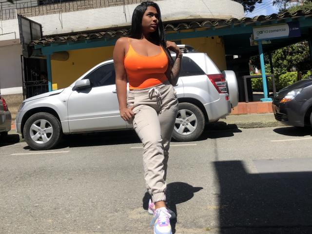 VickyGreys has dark skin, large boobs in an orange tank top, and wears beige pants with pink and white sneakers, standing in a parking lot with a white SUV.