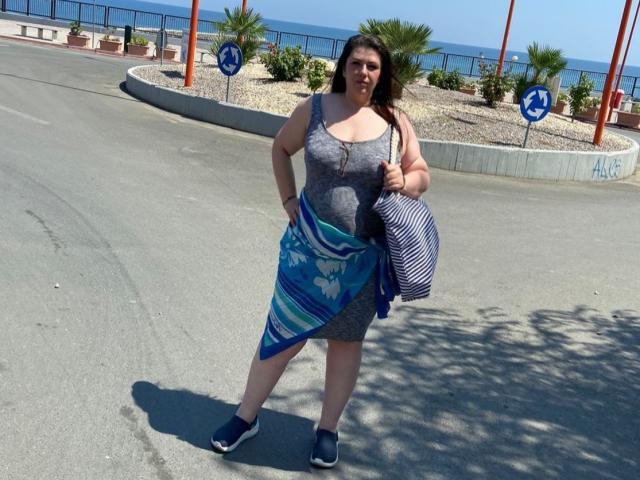 Carolina69, una mujer de talla grande con busto generoso, vestida con un vestido gris y zapatos azules, sosteniendo una toalla azul a rayas y un bolso a rayas, de pie en una calle soleada..