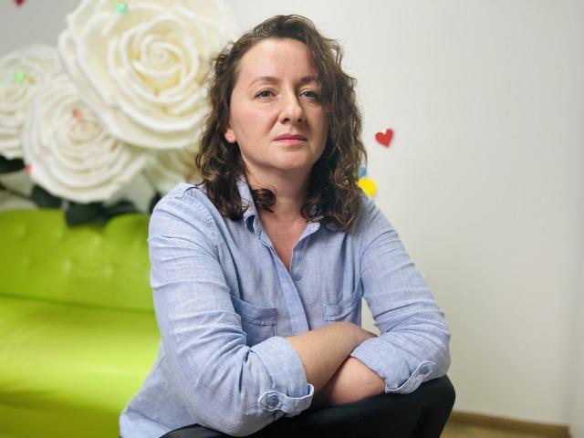 AbbyTaylorr has curly brown hair, a light blue shirt, and is sitting with arms crossed, looking slightly to the side with a subtle smile. Her pussy is not visible.
