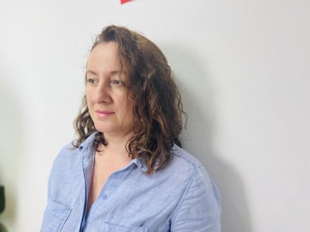 AbbyTaylorr has curly brown hair, light pink lipstick, and wears a blue button-up shirt, standing against a white wall with a slight smile.