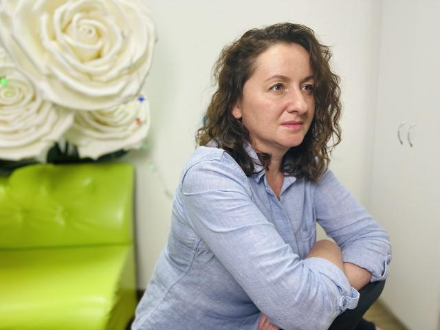 AbbyTaylorr with curly brown hair, light blue shirt, arms crossed, sitting, looking slightly to the side, green couch in background, large white rose wall decoration.