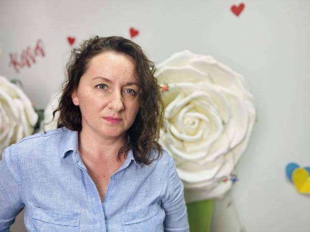 AbbyTaylorr with curly brown hair, wearing a light blue button-up shirt, has a serious expression, and large white rose decorations are in the background.