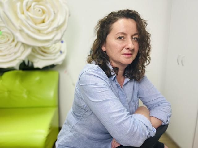 AbbyTaylorr, con i capelli castano ricci, indossa una camicia azzurra, seduta con le braccia conserte, guarda leggermente di lato, una sedia verde sullo sfondo, decorazione con una rosa bianca..