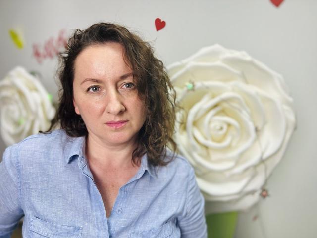 AbbyTaylorr with curly brown hair, wearing a blue button-up shirt, has a serious expression, and large white rose decorations are in the blurry background.