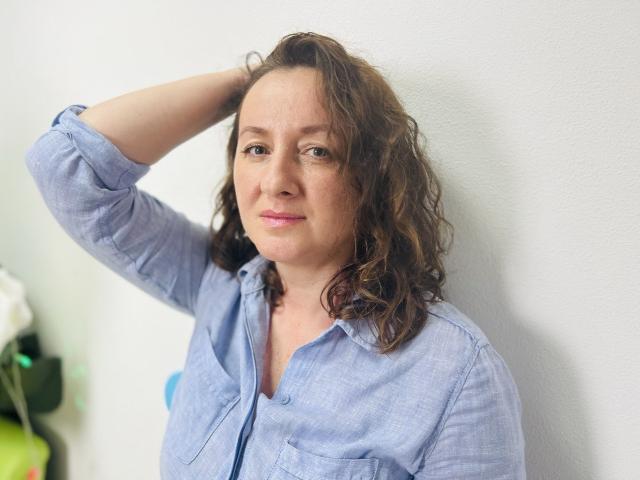 AbbyTaylorr with curly brown hair, wearing a blue button-up shirt, has her hand in her hair, showing a subtle smile, and has small, slightly visible tits.