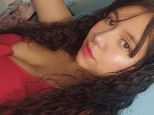 KrisGarner, com cabelo castanho encaracolado, piercing no nariz, blusa vermelha, lábios rosados e olhos castanhos, mostrando levemente os seios, tirando uma selfie..