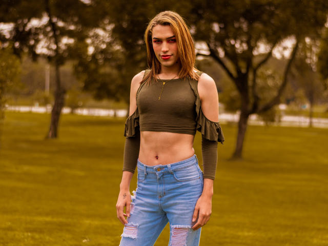 LaurynsKalii mit blonden Haaren, einem grünen, schulterfreien Crop-Top, das ihre Brüste zeigt, und zerrissenen blauen Jeans, steht draußen, wirkt selbstbewusst mit einem leichten Lächeln und trägt roten Lippenstift..