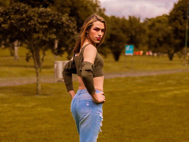 LaurynsKalii, con un corpo slanciato e tonico, indossa una canotta verde, jeans blu strappati, le mani sui fianchi, capelli castani, rossetto rosso, guarda indietro, in un parco erboso..