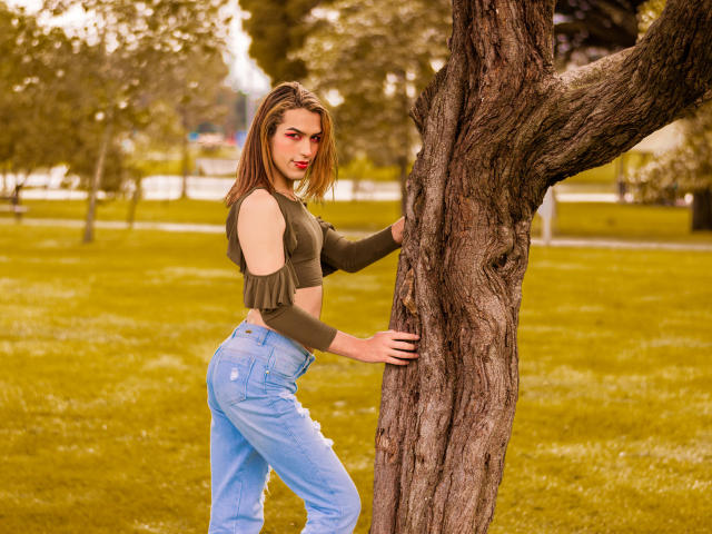 LaurynsKalii, com seios médios, uma blusa cropped verde e jeans azuis, está em pé contra uma árvore, olhando para a câmera, em um parque, com a mão direita apoiada no tronco..