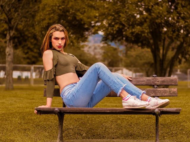 LaurynsKalii met een gemiddelde borstmaat, een groene crop top, blauwe jeans en witte sneakers, zittend op een houten bankje, benen gekruist, in een grasrijk park, kijkend naar de camera..