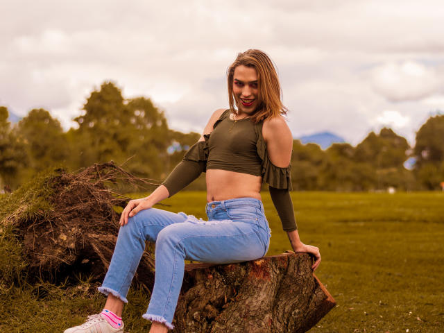 LaurynsKalii com seios médios, blusa cropped verde com um ombro de fora, jeans azuis e meias rosa, sentada em um toco de árvore, sorrindo, em um campo gramado, com céu nublado e árvores verdes ao fundo..