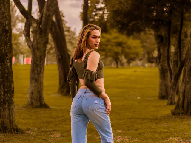 LaurynsKalii mit braunen Haaren, einem grünen Top mit einem Schulter frei, und blauen Jeans, steht in einem Park, blickt zurück und zeigt ihren Hintern, mit Bäumen im Hintergrund..