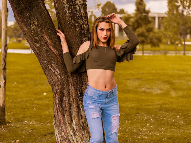 LaurynsKalii está em frente a uma árvore, vestindo uma blusa verde cropped com mangas que caem do ombro e jeans azuis rasgados, com a mão no cabelo, revelando seu abdômen plano e tonificado..