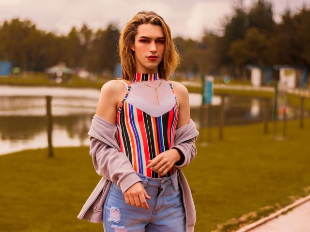 LaurynsKalii mit blonden Haaren, die ein farbenfrohes, gestreiftes Oberteil trägt, das einen Schulter frei lässt, blaue Jeans und eine graue Jacke, steht im Freien, zeigt ein leichtes, selbstbewusstes Lächeln und hat die Hände auf der Hüfte..