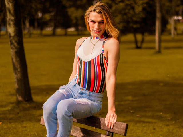 LaurynsKalii, com cabelo loiro, vestindo uma blusa colorida listrada e jeans rasgados, sentada em um banco de madeira, com os braços apoiados nos joelhos, olhando para a câmera, em um parque ensolarado..