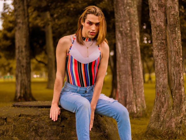 LaurynsKalii met blond haar dat tot op haar schouders reikt, gekleed in een gestreept tanktop en blauwe jeans, zittend op een boomstam in een bos, die lichtelijk opzij kijkt met een serieuze uitdrukking..