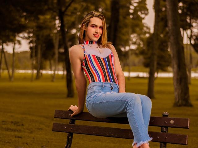 LaurynsKalii seduta su una panchina, indossa un top a tracolla colorato a righe e jeans blu, con le mani sulle ginocchia, in un parco con alberi. Il suo sedere è leggermente visibile..
