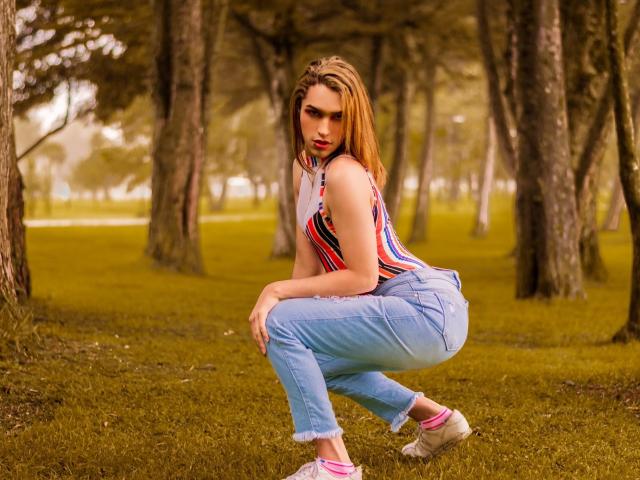LaurynsKalii hurkt in een park, gekleed in een gestreept mouwloos topje, blauwe jeans, roze sokken en witte schoenen. Ze heeft rode lippenstift en lang, golvend bruin haar..