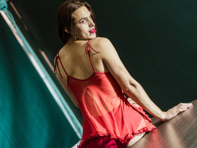 LaurynsKalii com lingerie vermelha transparente, de costas para a câmera, sentada em uma superfície de madeira, olhando por cima do ombro, cabelo castanho curto, batom vermelho, sexy, esguia, seios pequenos, fundo verde..
