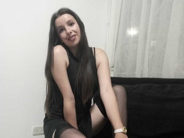 LebsGrup with long black hair, wearing a black dress and sheer black stockings, sitting on a black couch, smiling, showing her white wristwatch.