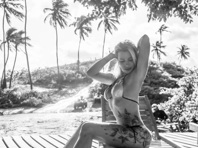 TatuadaSafadaHot with long blonde hair, bikini top, and floral tattoo on her ass, sitting on a wooden bench, hands in her hair, black and white, tropical setting.