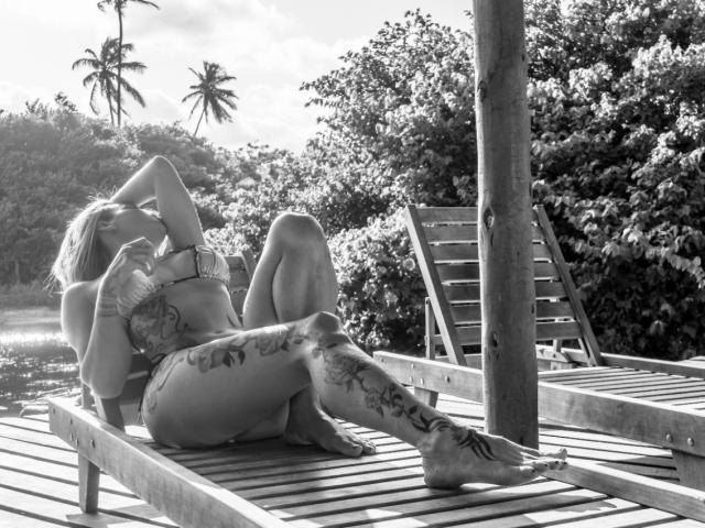 TatuadaSafadaHot with tattoos, lying on a wooden lounge chair, wearing a bikini, arms raised, legs spread, showing her ass and tattoos, in a black-and-white photo.
