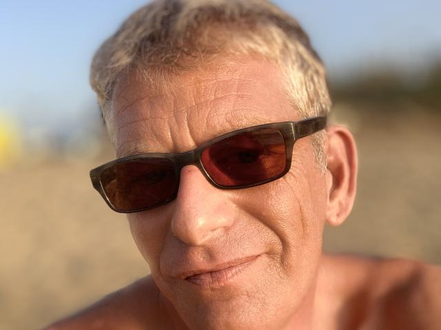 SergioEsp, shirtless, with blonde hair and brown sunglasses, has a slightly wrinkled, smiling face, and a tan, sunlit skin. His nose is small, and he looks relaxed.