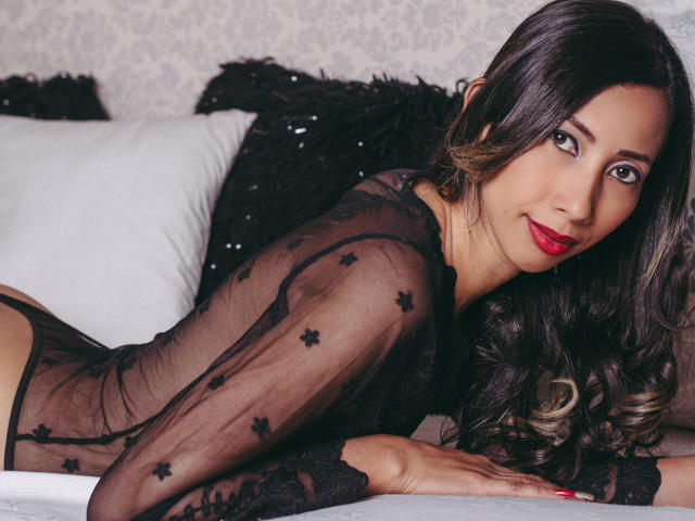 Isabelle Cruz, com cabelos longos e escuros, batom vermelho e blusa preta transparente, deitada em uma cama, mostrando suas nádegas, olhando para a câmera com um sorriso sedutor..