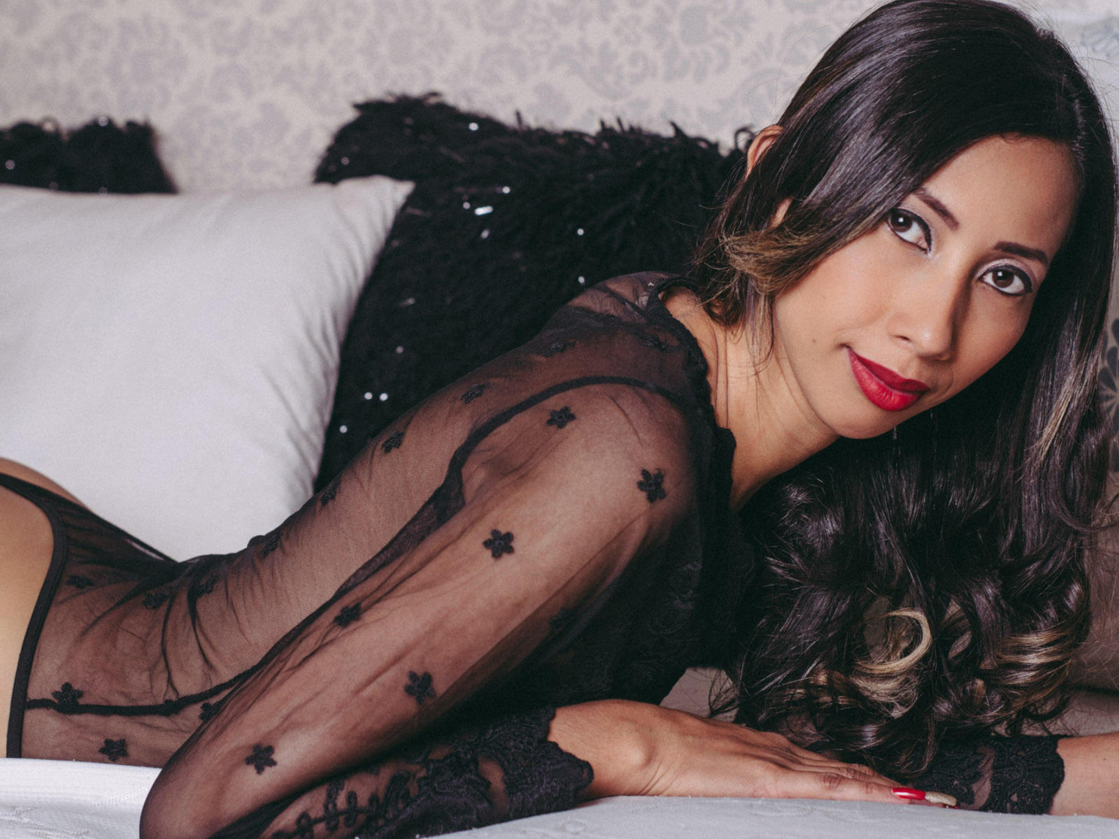 IsabelleCruz with long dark hair, red lipstick, and black sheer top, lying on a bed, showing her ass, looking at the camera with a sexy smile.