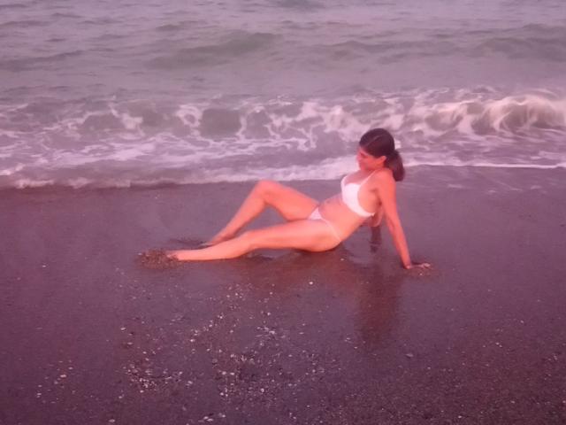 LaFrancaise with tan skin, dark hair in a ponytail, wearing a white bikini, sitting on wet black sand with small waves, legs spread, arms behind her, looking sideways.